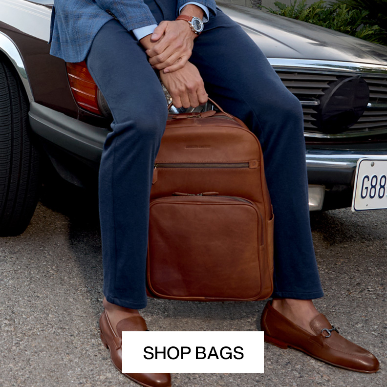 Man leaning on a vintage car holding onto a brown leather backpack with one hand. We see his blue colored pants and brown loafers.